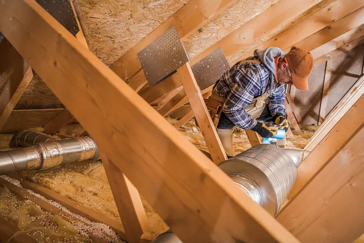A person wearing a plaid shirt, gloves, and a brown cap works in an attic. They are securing or inspecting silver ductwork among the wooden beams and insulation. The attic has a slanted roof and the person appears to be focused on their hvac service near me (crystal) task.
