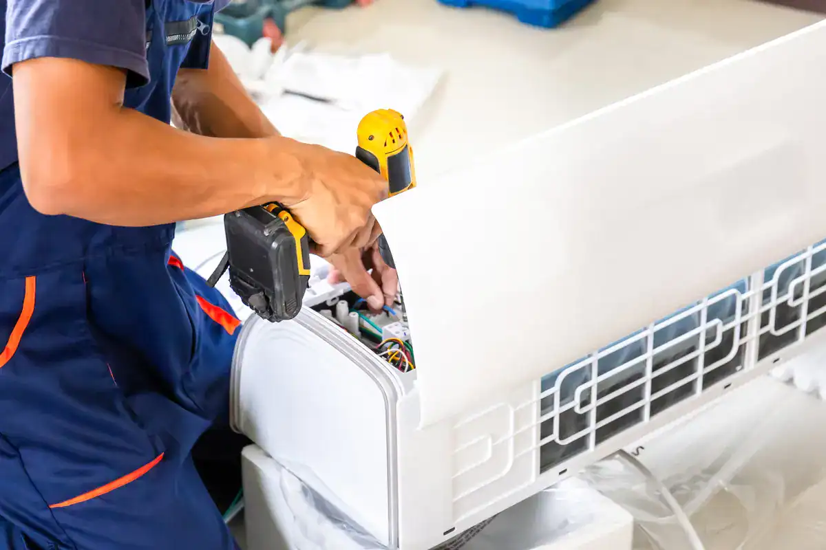 A technician in blue overalls is installing or repairing an air conditioning unit, using a yellow power drill. The inner components of the AC unit are exposed. The scene suggests a home or commercial repair service.