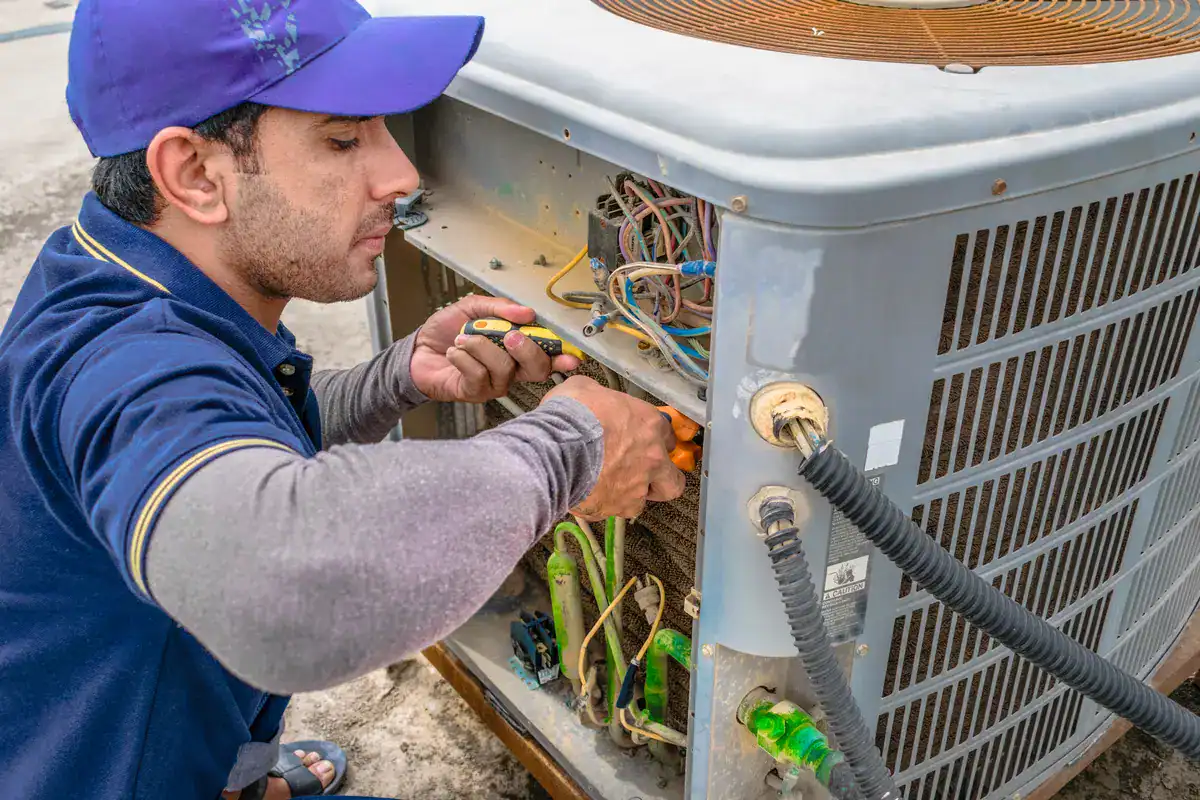 A technician in a blue cap and shirt uses tools to repair the wiring of an air conditioning unit on a rooftop. Various wires and components are visible as he works attentively on the exposed section of the unit—a reliable hvac service near me in Andover.