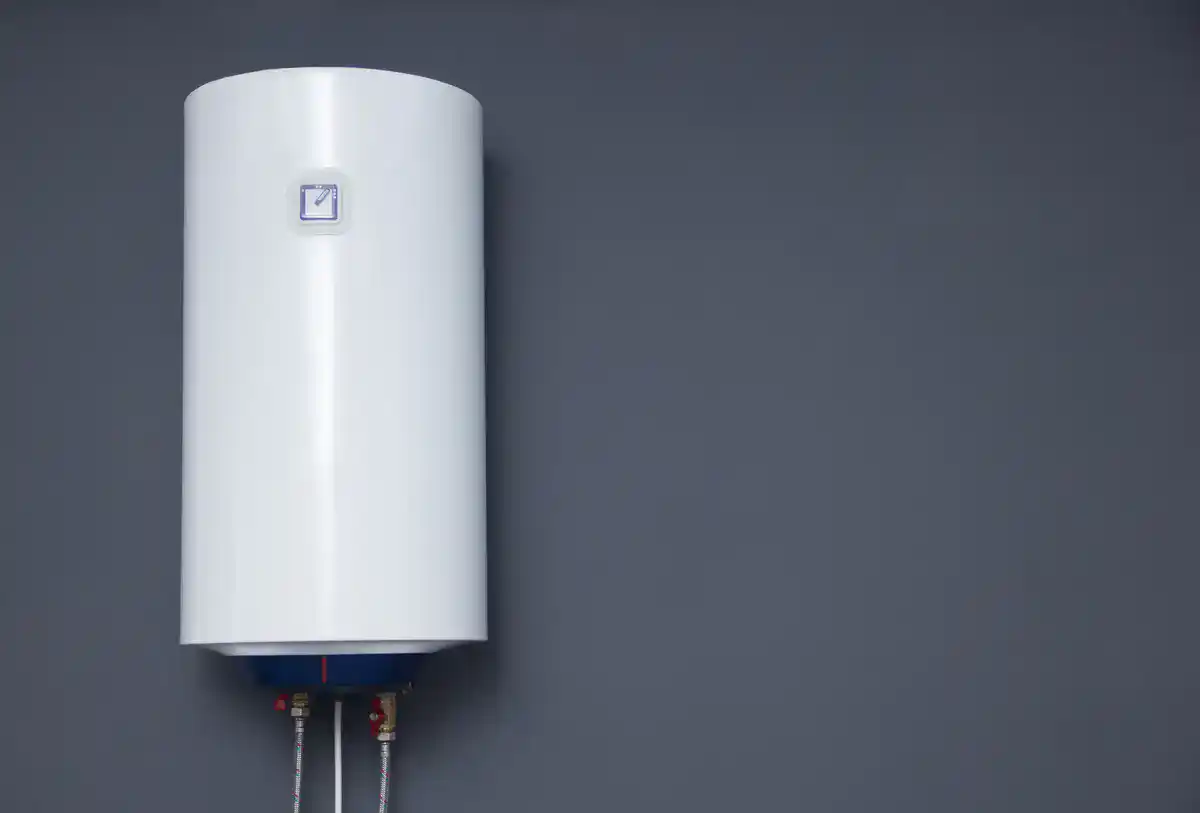 A white cylindrical water heater mounted on a dark gray wall, with visible pipes at the bottom. The unit has a small square display on the front near the top.