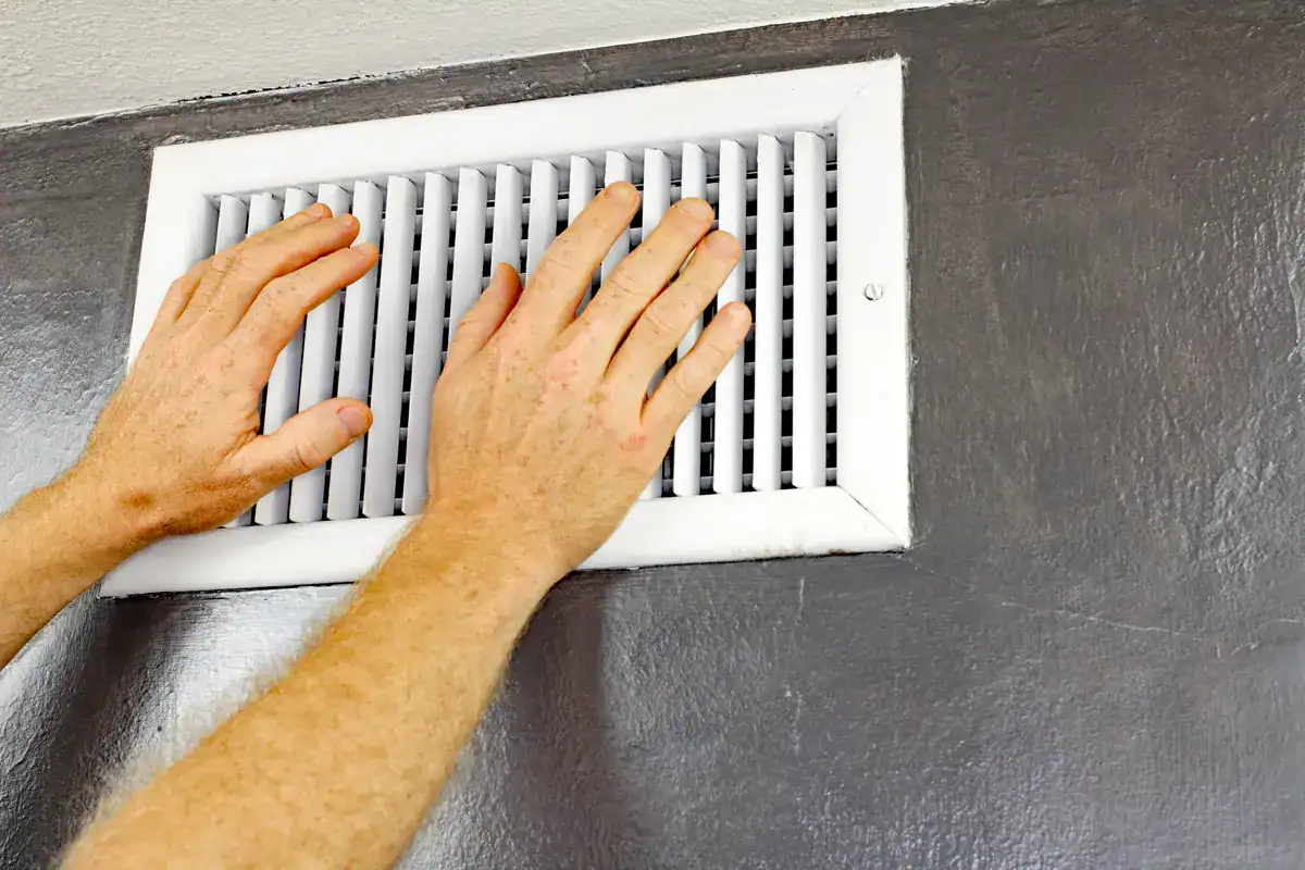 Two hands are placed on a white air vent on a gray wall, as if adjusting or feeling the airflow—a scene familiar to anyone searching for expert HVAC services near Woodbury.