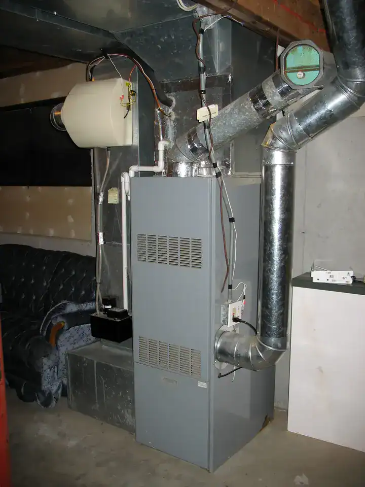 A basement with a large, gray furnace system connected to metal ducts and a humidifier mounted on the side. Some pipes and wiring are visible. There's an old couch in the background and a small white box on a nearby surface.
