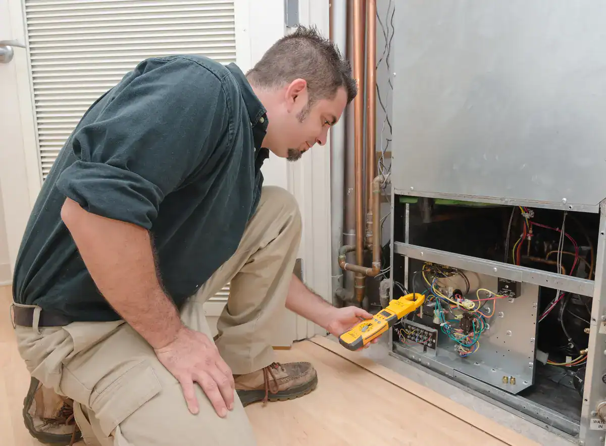 A technician kneels on the floor, inspecting HVAC equipment with a yellow digital multimeter. The open panel reveals various wires and components. Dressed in a dark shirt and light-colored pants, he embodies the expert care you'd expect from