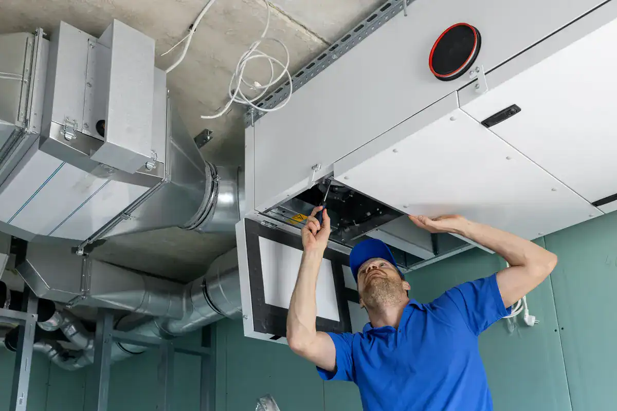 A technician in a blue uniform and cap is expertly using a screwdriver on a ceiling-mounted air conditioning unit. Surrounded by various ducts and wires, the setting hints at an industrial vibe, typical of HVAC tasks in commercial spaces near Edina.