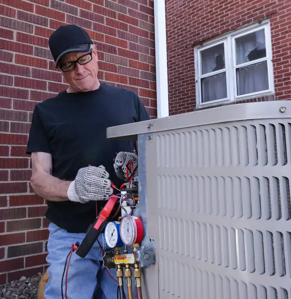 A person in a black cap and shirt, wearing gloves, is using tools to repair or inspect an air conditioning unit outside a brick building. They are working with gauges attached to the unit.