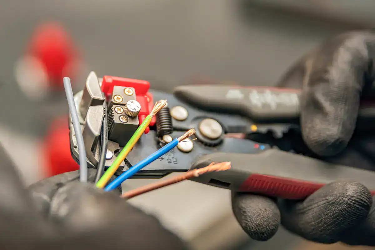 A close-up of hands in black gloves using wire strippers to skillfully strip insulation from electrical wires, handled with the expertise you'd find from an electrician near me in Oshkosh. The tools grip multicolored wires—green, yellow, and blue—set against a softly blurred background.