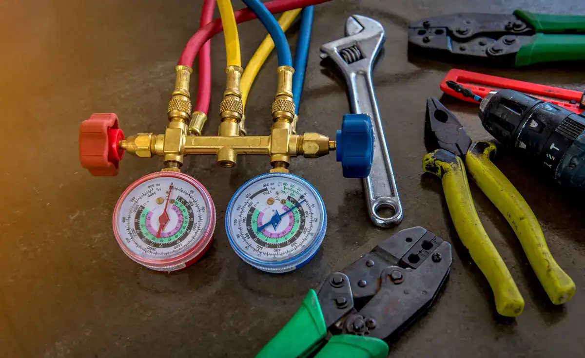 A set of HVAC gauges with red, blue, and yellow hoses is placed on a concrete surface. Nearby are assorted tools: a wrench, wire cutters, a drill, and crimping pliers with green handles.