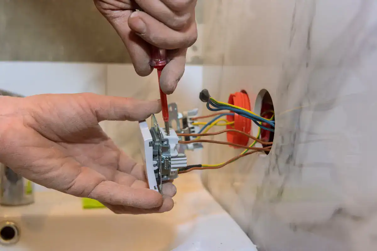 A person, possibly an electrician near Green Bay, installs an electrical outlet in a wall. One hand holds the outlet while the other uses a screwdriver to secure it. Colorful wires peek from the wall opening against a backdrop of tiles and a partially visible countertop.