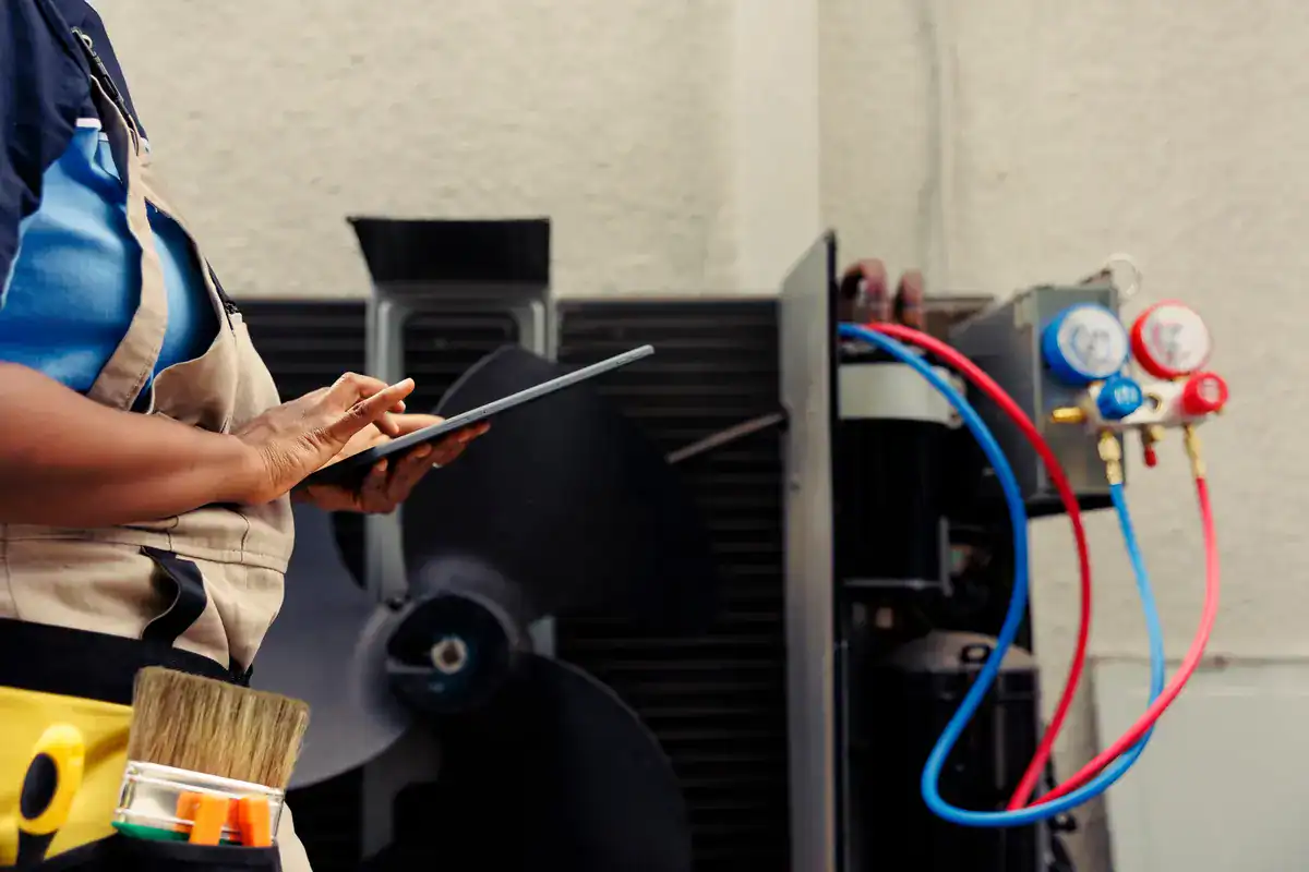 A person wearing a work apron holds a tablet while standing in front of HVAC equipment. Various tools are visible in their apron, and gauges with red and blue hoses are connected to the equipment. They seem ready for any HVAC need, perhaps even for those searching 