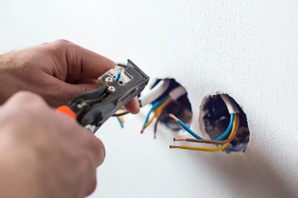 Close-up of an electrician near Ashwaubenon using pliers to work on electrical wires protruding from two small openings in a white wall. The wires, in blue, yellow-green, and brown hues, hint at ongoing installation or repair.