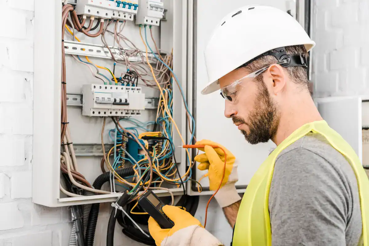 An electrician near me in Sturgeon Bay wearing a safety vest, helmet, and gloves uses a multimeter to test electrical connections in an open panel. Wires and circuit breakers are visible inside the panel on a white brick wall.