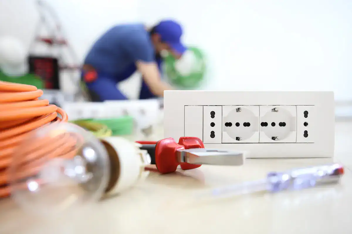 Close-up of a double electrical outlet on a table beside tools like a screwdriver and a bundle of orange cable. In the background, an electrician in blue work attire is blurred, handling equipment, indicating the presence of skilled professionals available for those searching