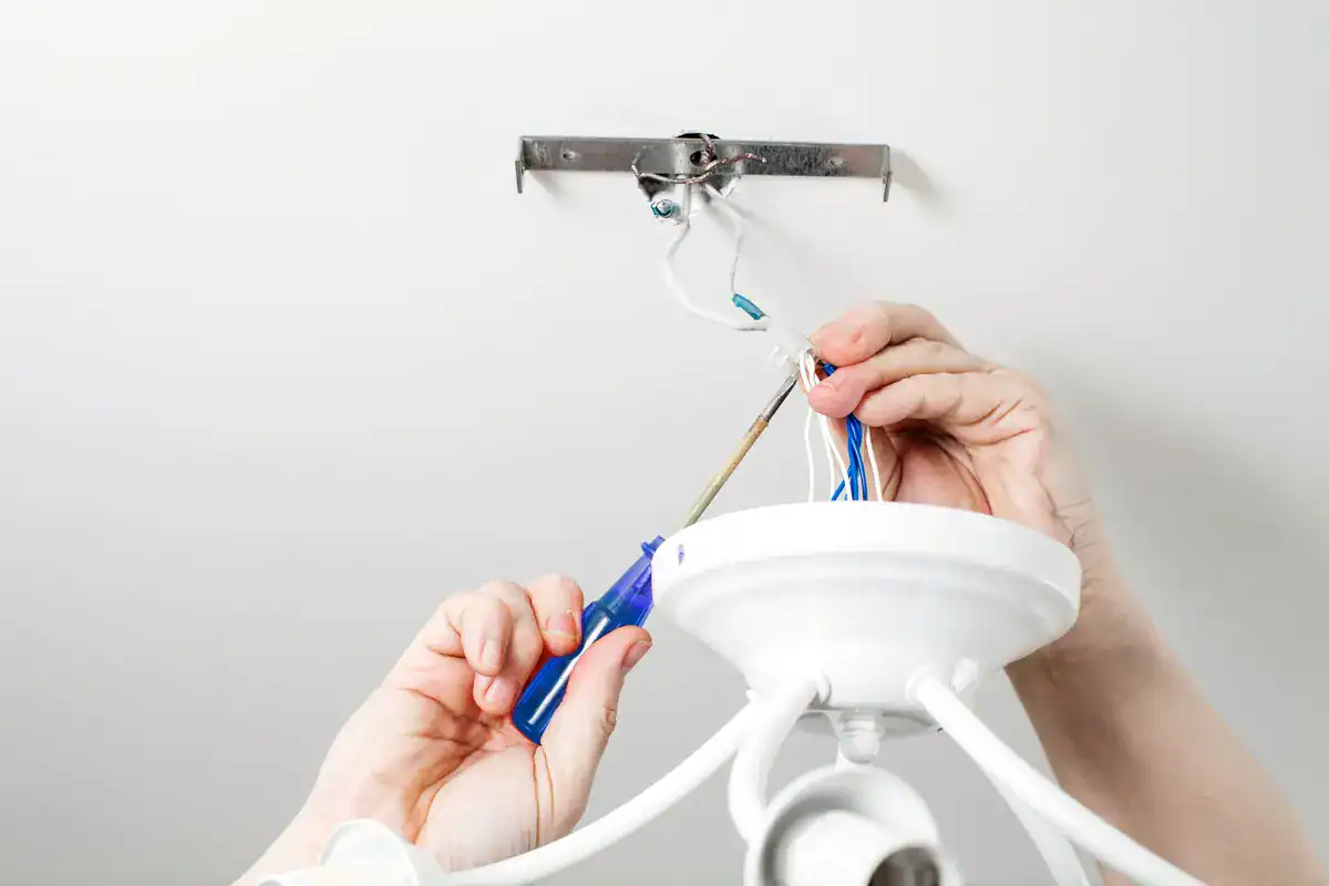 An electrician near me in Neenah skillfully installs a white ceiling light fixture, using a blue screwdriver to connect wires. Hands firmly grip both the fixture and tool against the white ceiling, with exposed wires visible.