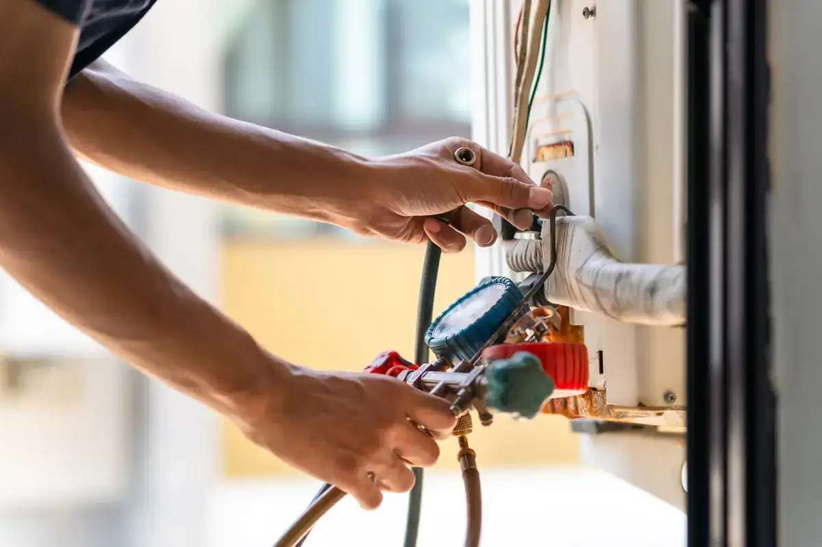 A technician carefully adjusts connections on an air conditioning unit, using gauges and hoses for maintenance. The focus is on skilled hands and precise tools, with part of the HVAC system visible in the background. For those seeking 