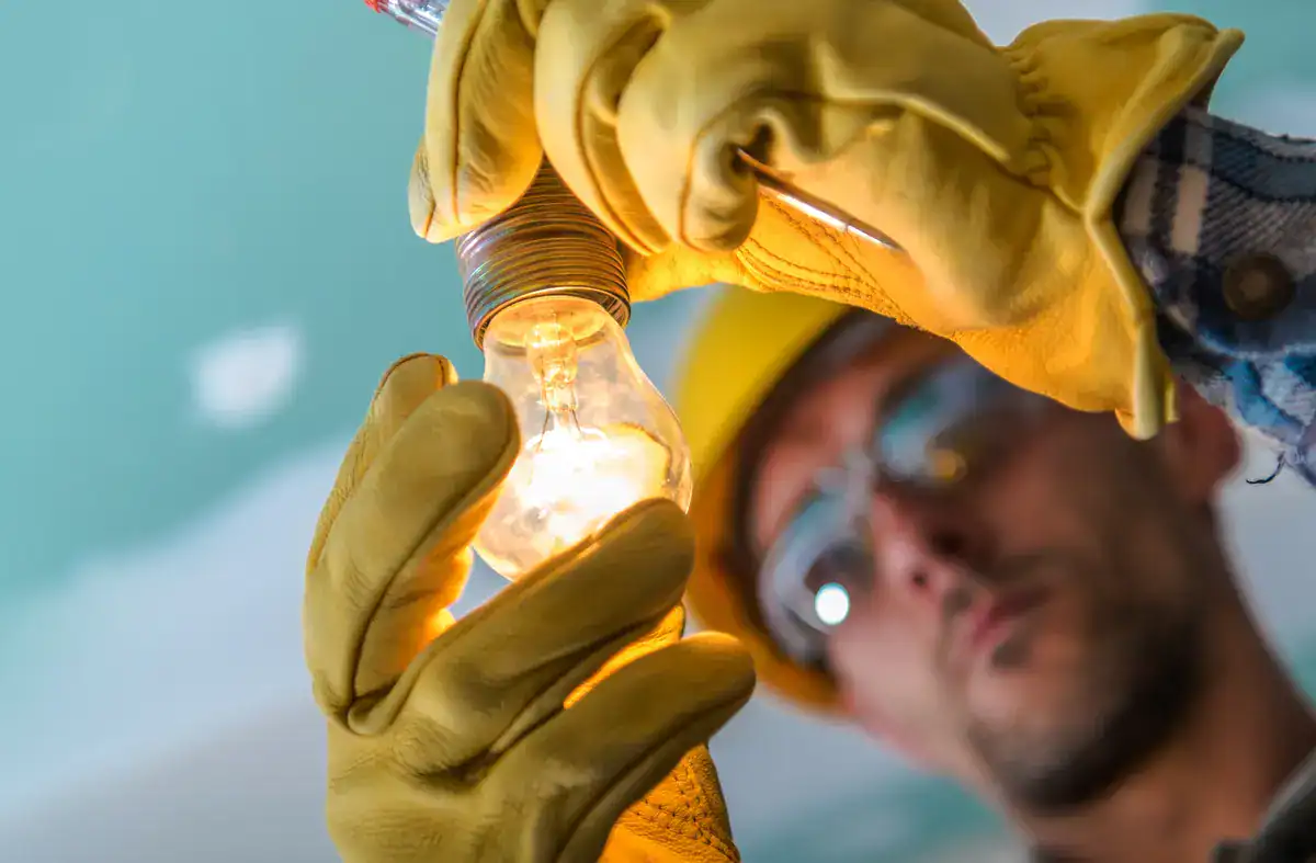 A person wearing gloves and a hard hat, likely an electrician near me in Menasha, installs a glowing light bulb into a ceiling fixture. The background is slightly blurred, emphasizing the skillful hands and the bulb against a light green ceiling with visible patches.