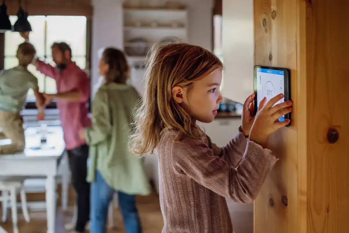 A young girl adjusts a smart thermostat on a wooden wall, optimizing the family's comfort with an HVAC system. In the background, her family gathers around a warmly lit kitchen table in their Menasha home, where cozy shelves complete the inviting atmosphere.