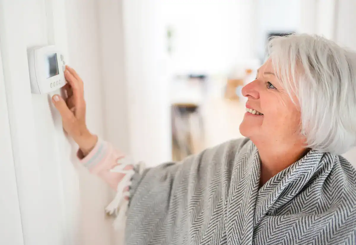 A smiling elderly woman with short white hair adjusts a thermostat on the wall, contemplating the efficiency of her hvac system in Oshkosh. Wrapped in a patterned gray shawl, she stands comfortably in her bright room.