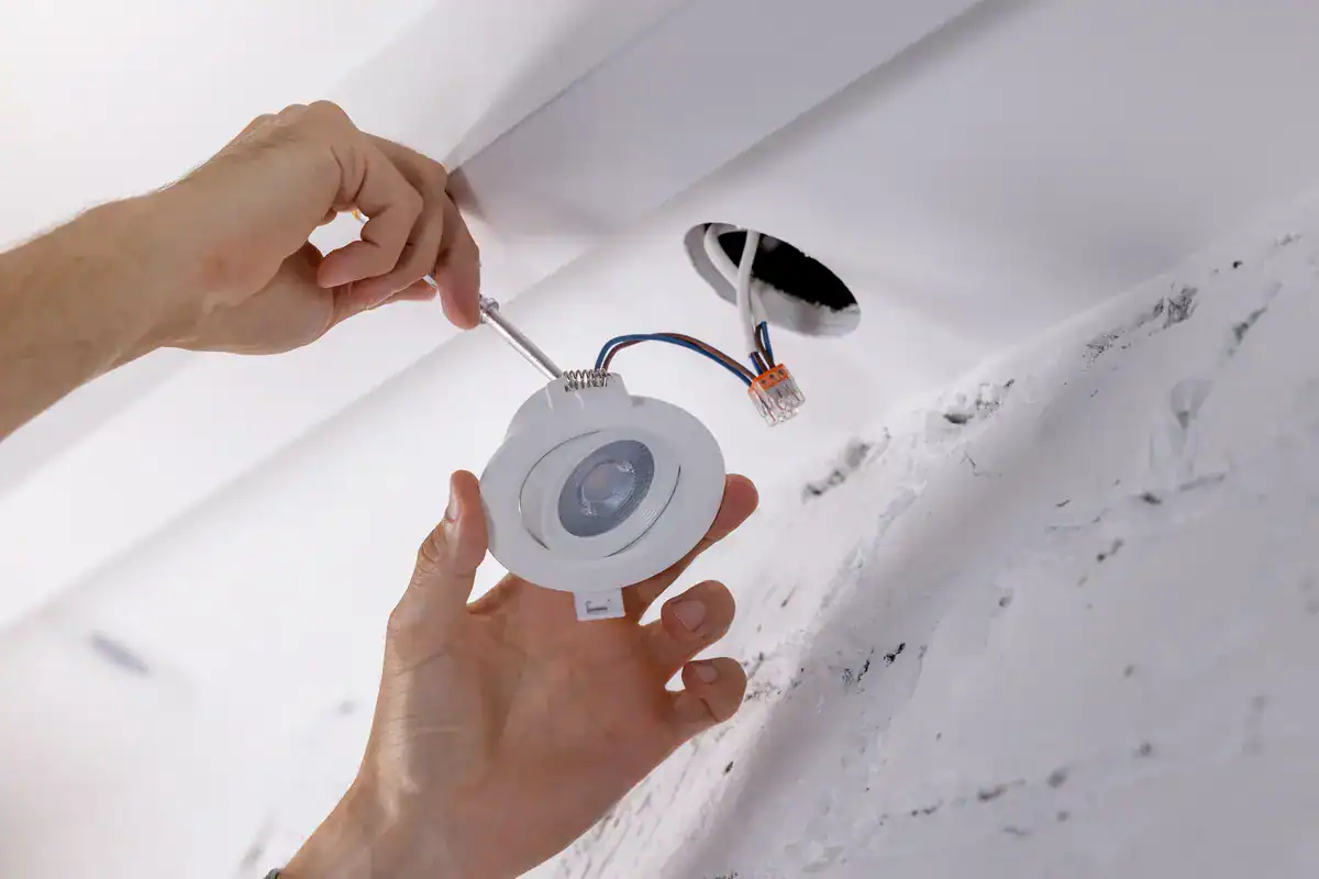 Two hands are carefully installing a recessed ceiling light fixture, expertly connecting wires before placing it into the ceiling's opening. The white plaster shows signs of recent construction, making it clear that an electrician near me in New London has been hard at work.