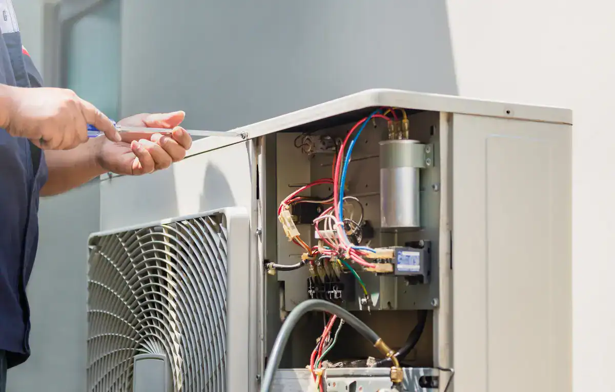 A technician repairs an outdoor HVAC unit, deftly using a screwdriver to adjust components. The open panel reveals colorful wires and internal parts, as the local Allouez sun casts natural light on the skilled hands maintaining the system.