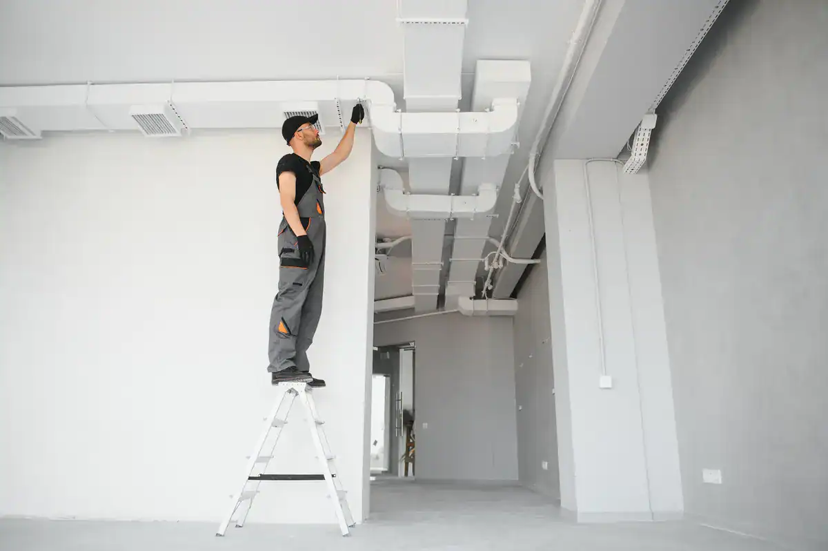 A worker in overalls and a hat stands on a ladder, inspecting ductwork on the ceiling of an unfinished room. The space, reminiscent of many HVAC projects in De Pere, has exposed ventilation and minimal decor, featuring gray walls and a concrete floor.