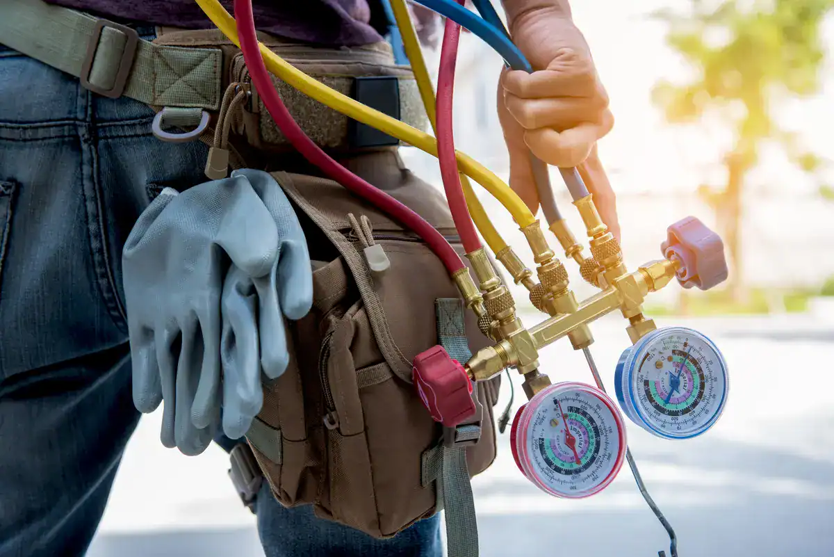 A person holds a set of HVAC gauges with red, yellow, and blue hoses, alongside a tool bag with attached work gloves, standing outside in daylight. The focus on equipment hints at air conditioning or refrigeration tasks—an ideal scene for those seeking 