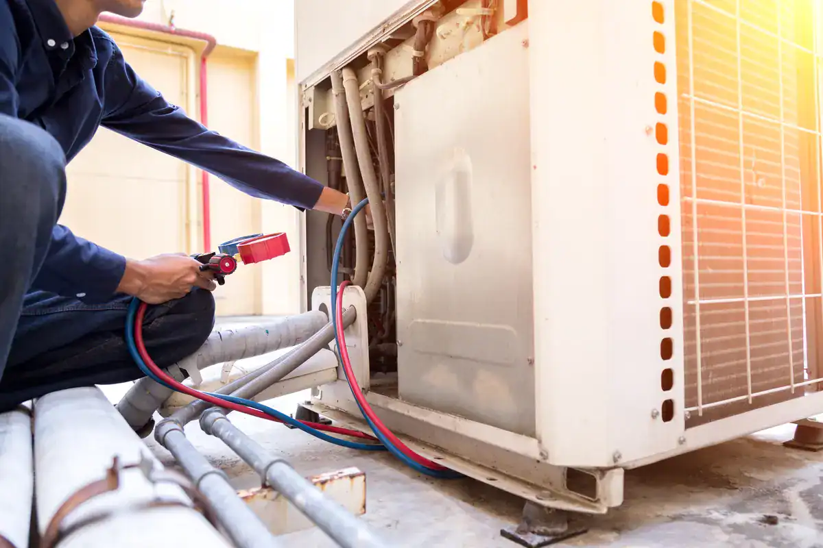 A technician is kneeling while examining a large HVAC unit. They are holding gauges connected by hoses to the unit, with sunlight streaming onto the equipment. The scene suggests maintenance or repair work on the air conditioning system.