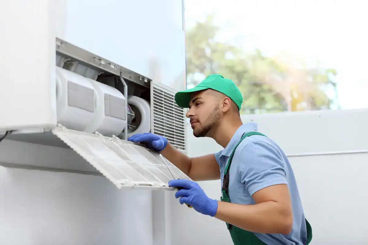 A technician in a green cap and blue gloves examines an air conditioning unit. He holds a removed filter panel, inspecting the components inside. The background shows blurred greenery through a window, suggesting an indoor setting.