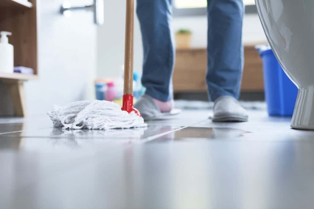 A person mops a tiled floor in a bathroom. The image shows the mop head on the tiles, with the person's legs in slippers visible. In the background, there's a blue bucket and a wooden cabinet.