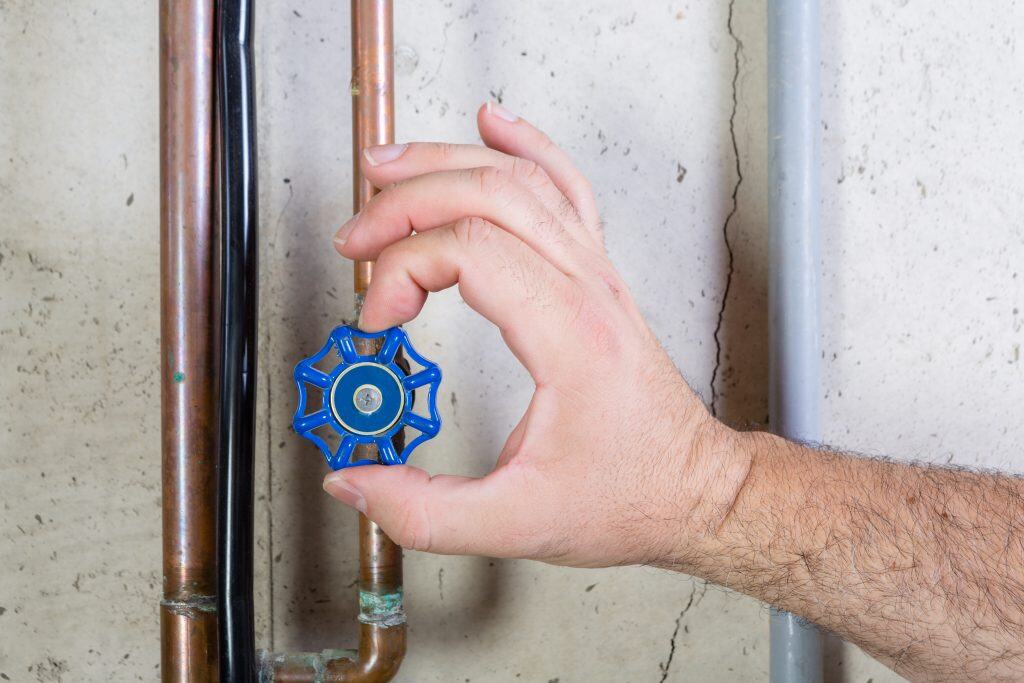 A hand is turning a blue valve on a copper pipe against a concrete wall. Two pipes run parallel to each other, one copper and one gray. The valve is shaped like a gear. The wall is rough with visible lines and cracks.
