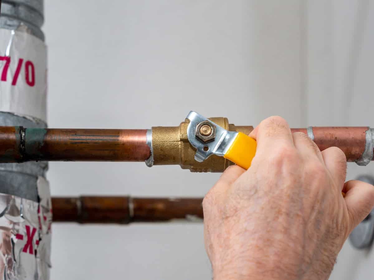 A hand using a pipe cutter to cut a copper pipe. The cutter has a yellow handle and is positioned around the pipe, which is connected to a brass valve. The background is a plain wall.