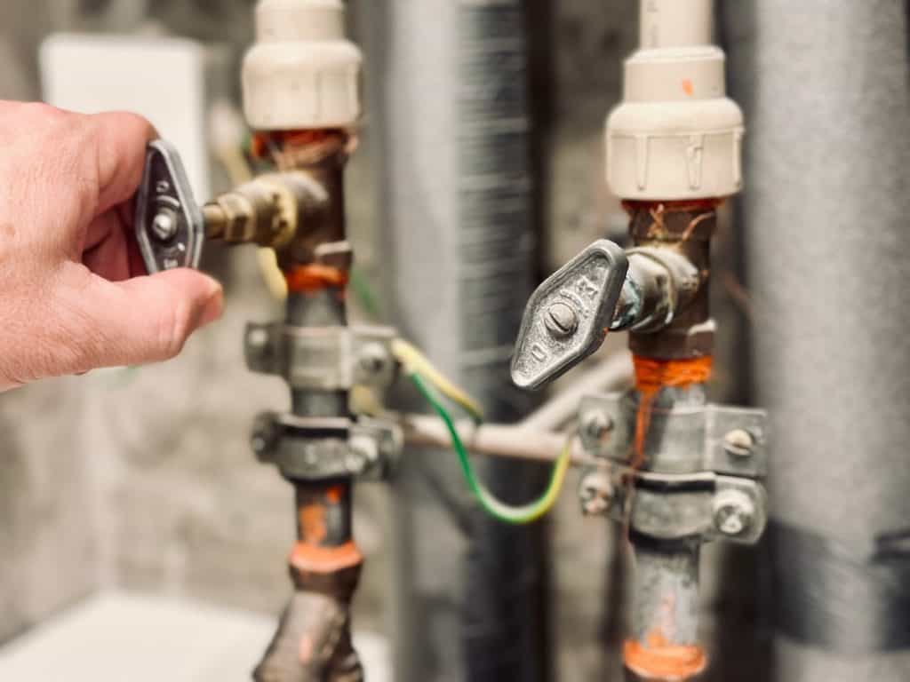 A hand turning one of two angled metal valves attached to pipes. The valves have orange markings, and the pipes are surrounded by insulation. The setting appears to be an industrial or utility area.