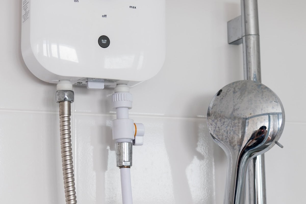 Close-up of a white electric water heater attached to a wall, with a metallic shower head connected by a flexible hose. White tiles form the backdrop, and there are visible control knobs on the heater.