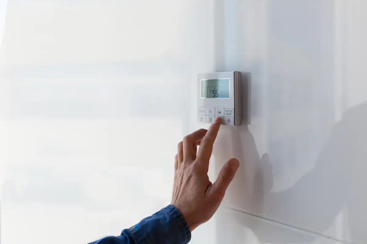 A hand adjusting a digital thermostat mounted on a white wall. The display shows a temperature setting. The person wears a denim shirt, and their finger is poised over a button on the thermostat.