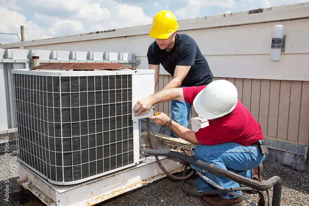 Two workers in hard hats are repairing or installing an HVAC unit on a rooftop. One person is wearing a black shirt and yellow hard hat, the other a red shirt and white hard hat. They appear focused on their task, with tools in use.