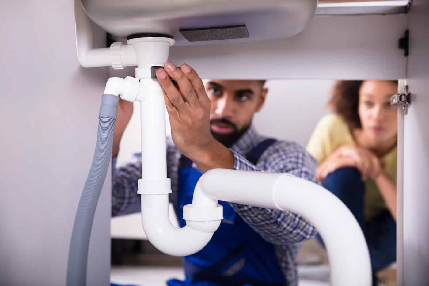 A plumber in a blue uniform is expertly fixing the pipes under a sink, offering invaluable tips on drain cleaning near me in Lakeville, MN. A person with curly hair watches attentively from the side. Several white and gray pipes are visible, with the focus on the plumber's hands.