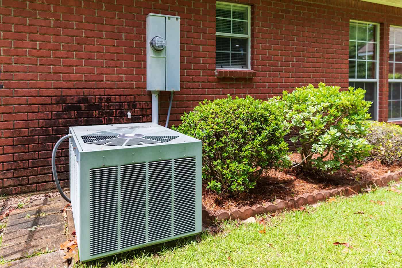 Older Style Air Conditioner System Next To Home With Brick, Bushes And Clean Yard.